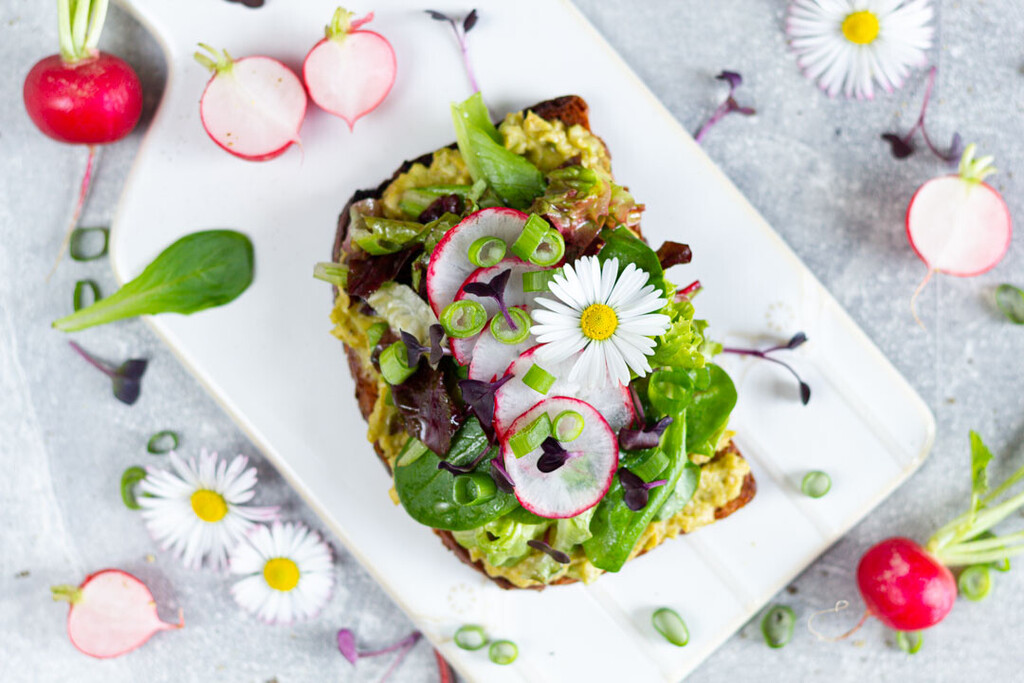 Nahaufnahme einer belegten Avocado-Stulle mit Radieschenscheiben, Salat und Frühlingszwiebeln auf dunklem Brot.