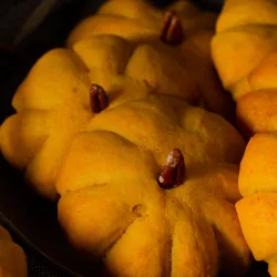 Fluffige Kürbisbrötchen Rezept: Das perfekte Rezept für dein Halloween Gebäck Kürbisbrötchen Rezept: Goldgelb gebackene Teiglinge in Kürbisform mit Pecannuss-Stiel auf einem dunklen Teller.