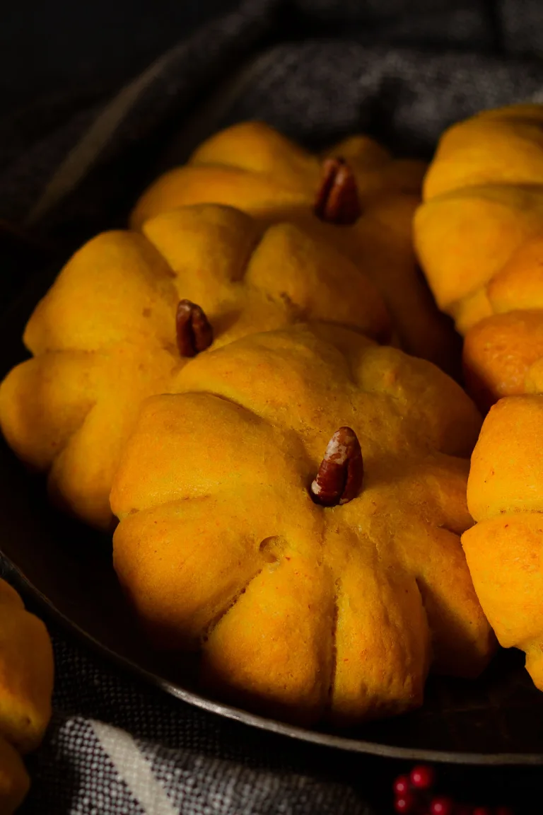 Kürbisbrötchen Rezept: Goldgelb gebackene Teiglinge in Kürbisform mit Pecannuss-Stiel auf einem dunklen Teller.