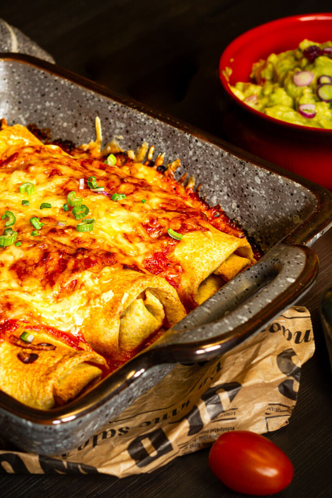 Frisch gebackene vegetarische Enchiladas in einer Auflaufform, mit geschmolzenem Käse und einer roten Sauce überbacken, garniert mit frischen Kräutern.