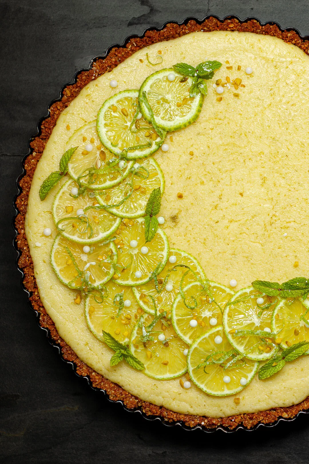 Cremiges Key Lime Pie Rezept mit knusprigem braunem Boden, dekoriert mit frischen Limettenscheiben, Minzblättern und weißen Zuckerperlen auf dunklem Untergrund.