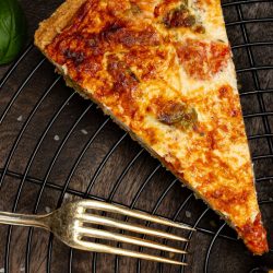 Goldbraun gebackene Quiche mit gerösteten Tomaten, Basilikum und Parmesan, ein Stück herausgeschnitten, dekoriert mit frischem Basilikum und Cherrytomaten