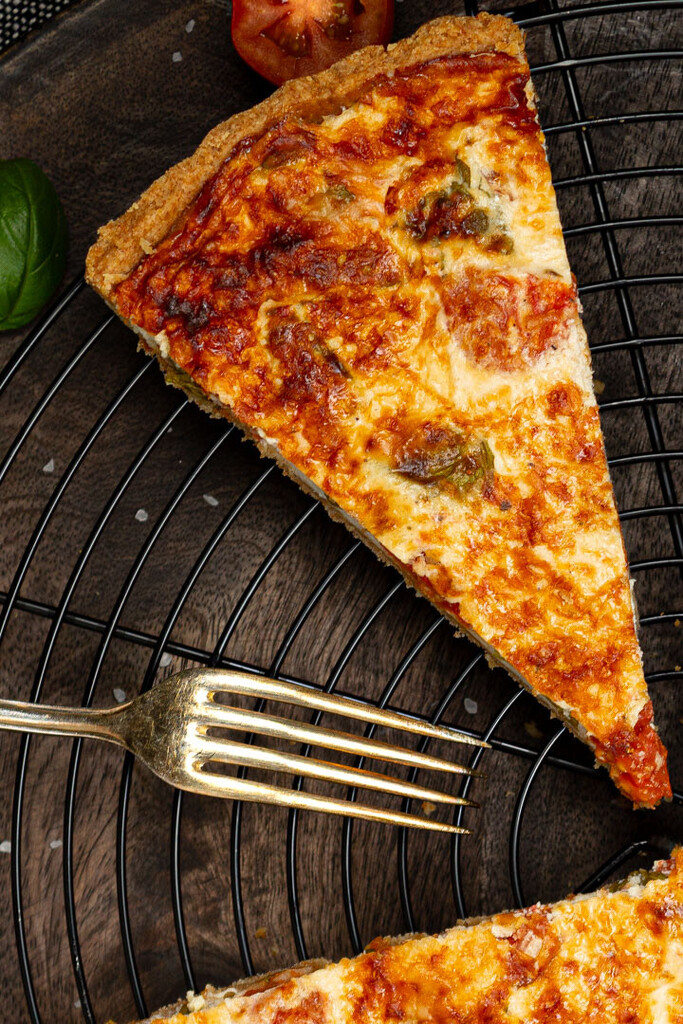 Goldbraun gebackene Quiche mit gerösteten Tomaten, Basilikum und Parmesan, ein Stück herausgeschnitten, dekoriert mit frischem Basilikum und Cherrytomaten