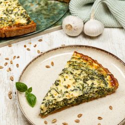 Ein Stück Spinat-Quiche mit goldbrauner Oberfläche auf einem Teller, im Hintergrund eine ganze Quiche sowie Knoblauch und frische Kräuter dekoriert