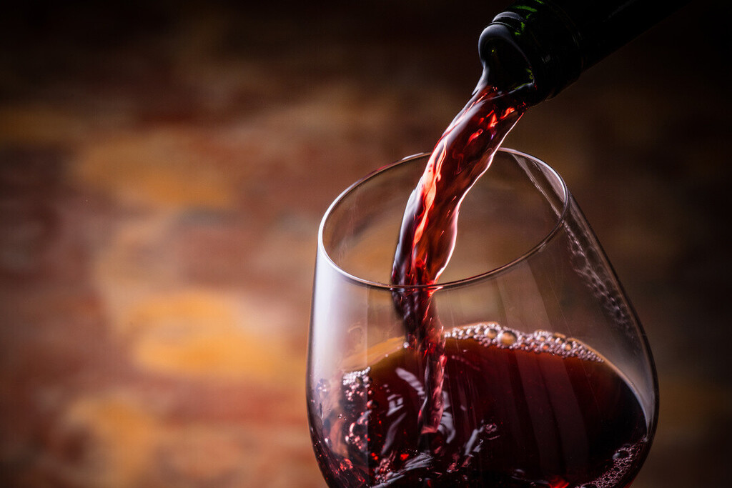 Grundlagen der Weinkunde: Dunkelroter Rotwein wird aus einer Flasche in ein bauchiges Weinglas vor warmem, unscharfem Hintergrund eingeschenkt.