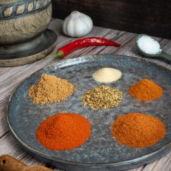 Verschiedene gemahlene mexikanische Gewürze wie Paprika, Kreuzkümmel und Chili, die dekorativ in kleinen Häufchen und in einem Glasgefäß auf einem dunklen Untergrund angeordnet sind.