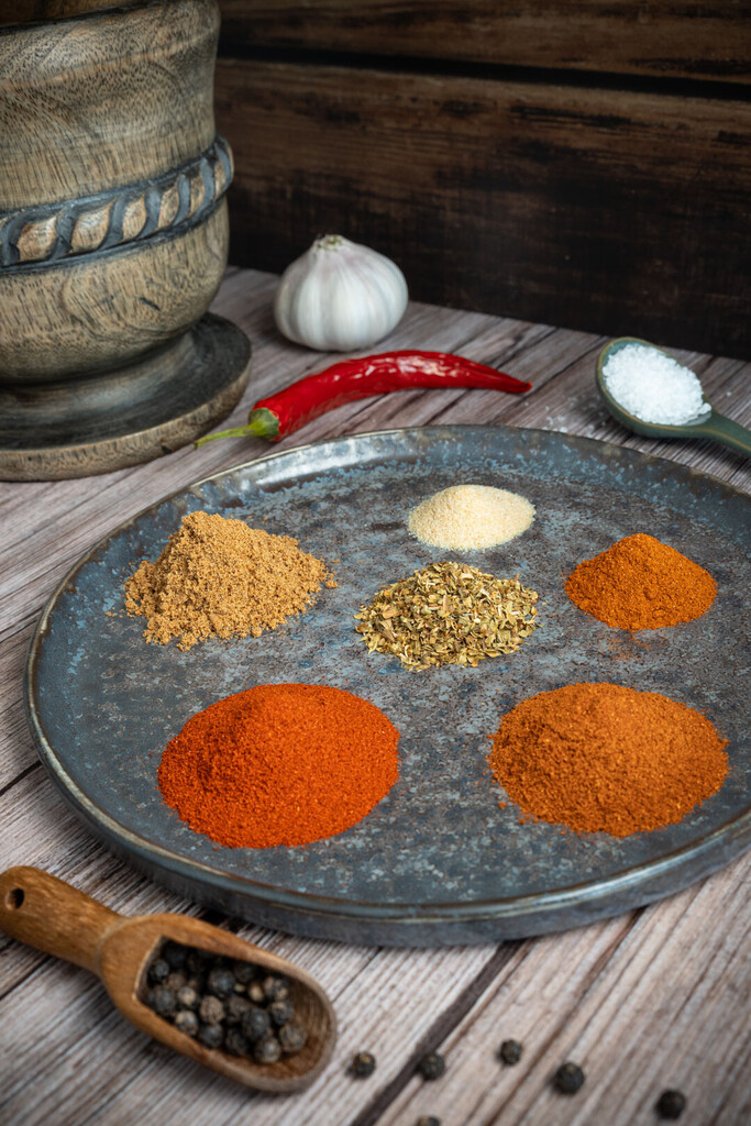 Verschiedene gemahlene mexikanische Gewürze wie Paprika, Kreuzkümmel und Chili, die dekorativ in kleinen Häufchen und in einem Glasgefäß auf einem dunklen Untergrund angeordnet sind.