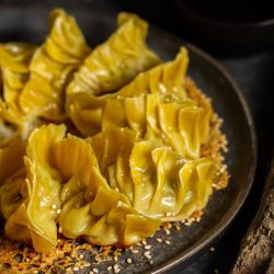 Goldbraun angebratene japanische Gyoza-Teigtaschen auf einem dunklen Keramikteller, bestreut mit Sesam. Im Hintergrund sind Essstäbchen und eine kleine Schale mit Sauce zu sehen.