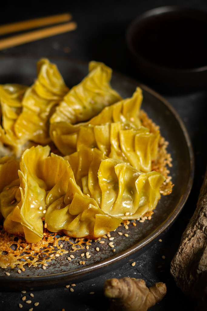 Goldbraun angebratene japanische Gyoza-Teigtaschen auf einem dunklen Keramikteller, bestreut mit Sesam. Im Hintergrund sind Essstäbchen und eine kleine Schale mit Sauce zu sehen.