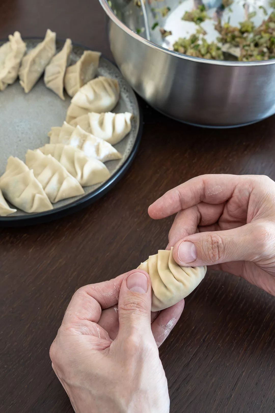 Hände falten einen gefüllten Gyoza mit typischen Falten, im Hintergrund liegen weitere rohe japanische Teigtaschen auf einem Teller.