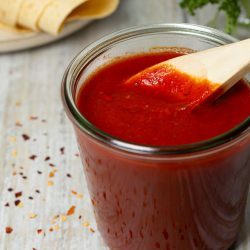 Beste Enchilada Sauce: Ohne Konservierungsstoffe & super würzig Rote, sämige Enchilada-Sauce in einem kleinen Glasbehälter, umgeben von mexikanischen Gewürzen und frischen Zutaten auf einem dunklen Hintergrund.