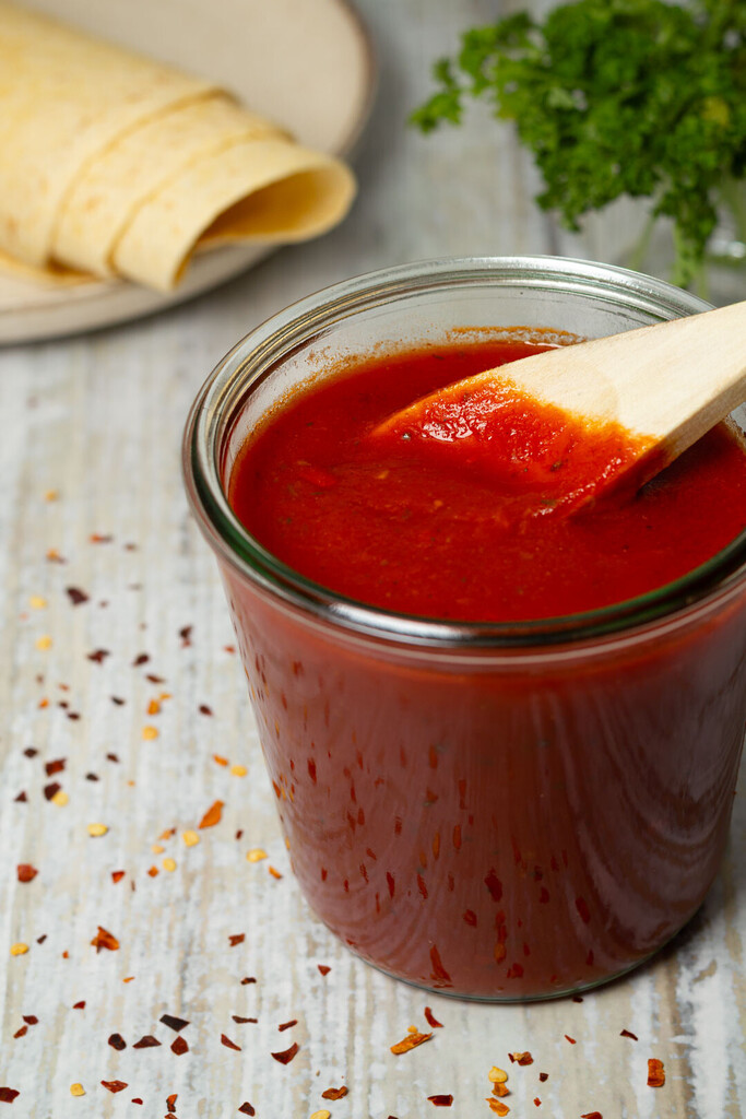 Rote, sämige Enchilada-Sauce in einem kleinen Glasbehälter, umgeben von mexikanischen Gewürzen und frischen Zutaten auf einem dunklen Hintergrund.