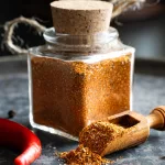 Mexikanische Gewürzmischung selber machen: Mexikanische Gewürzmischung in einem Gewürzglas, umgeben von ganzen Chilis und Pfefferkörnern auf Stein.