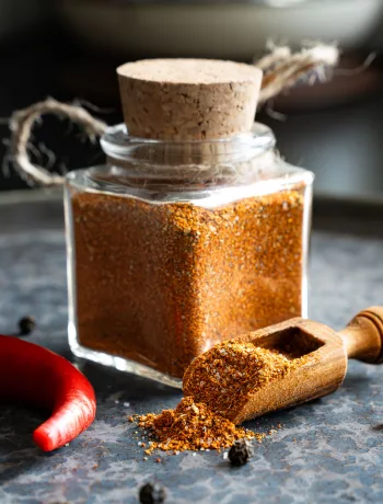 Mexikanische Gewürzmischung selber machen: Mexikanische Gewürzmischung in einem Gewürzglas, umgeben von ganzen Chilis und Pfefferkörnern auf Stein.