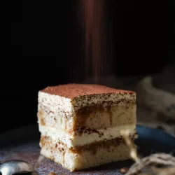 Tiramisu ohne Alkohol und Ei: Cremiges italienisches Rezept Ein Stück italienisches Tiramisu ohne Alkohol und Ei mit sichtbaren Biskuit- und Mascarponeschichten auf einem dunklen Teller, während es von oben mit Kakaopulver bestäubt wird.