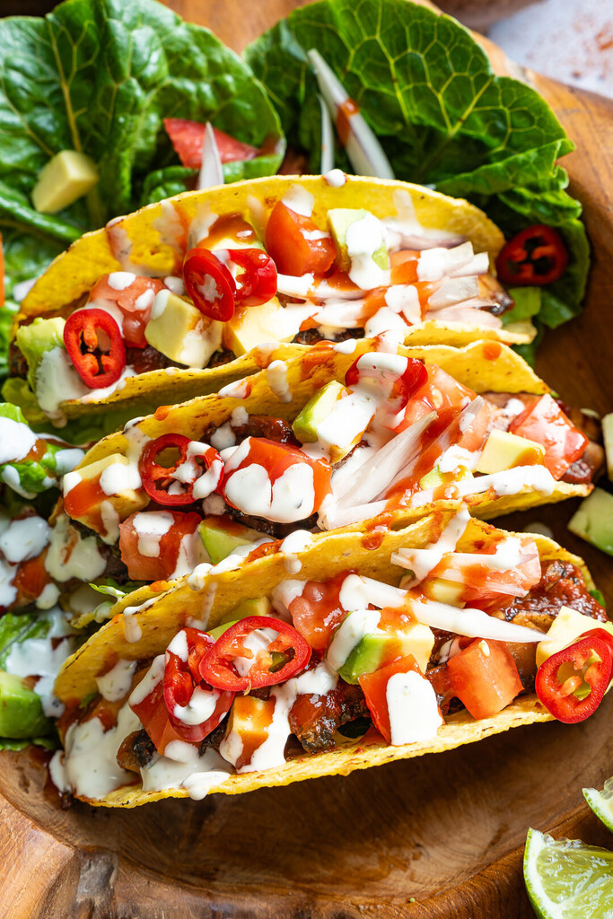 Drei bunte mexikanische Tacos in weichen Tortillas, gefüllt mit gebratenen Portobello-Pilzen, cremiger Avocado, frischen Tomaten und einer feinen Sauce, serviert auf einem Holzbrett.