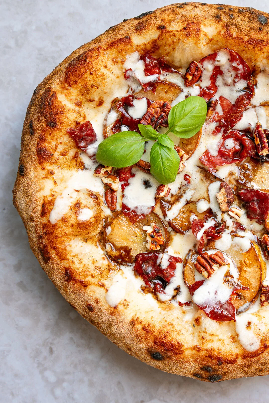 Draufsicht auf eine hausgemachte Pizza Bianca mit einem hohen, luftig-gebräunten Rand im neapolitanischen Stil. Der Belag besteht aus geschmolzenem Käse, hauchdünnem Bresaola, goldbraun karamellisierten Birnenscheiben und gerösteten Pekannüssen. Ein frischer Basilikumzweig ziert die Mitte der Pizza.