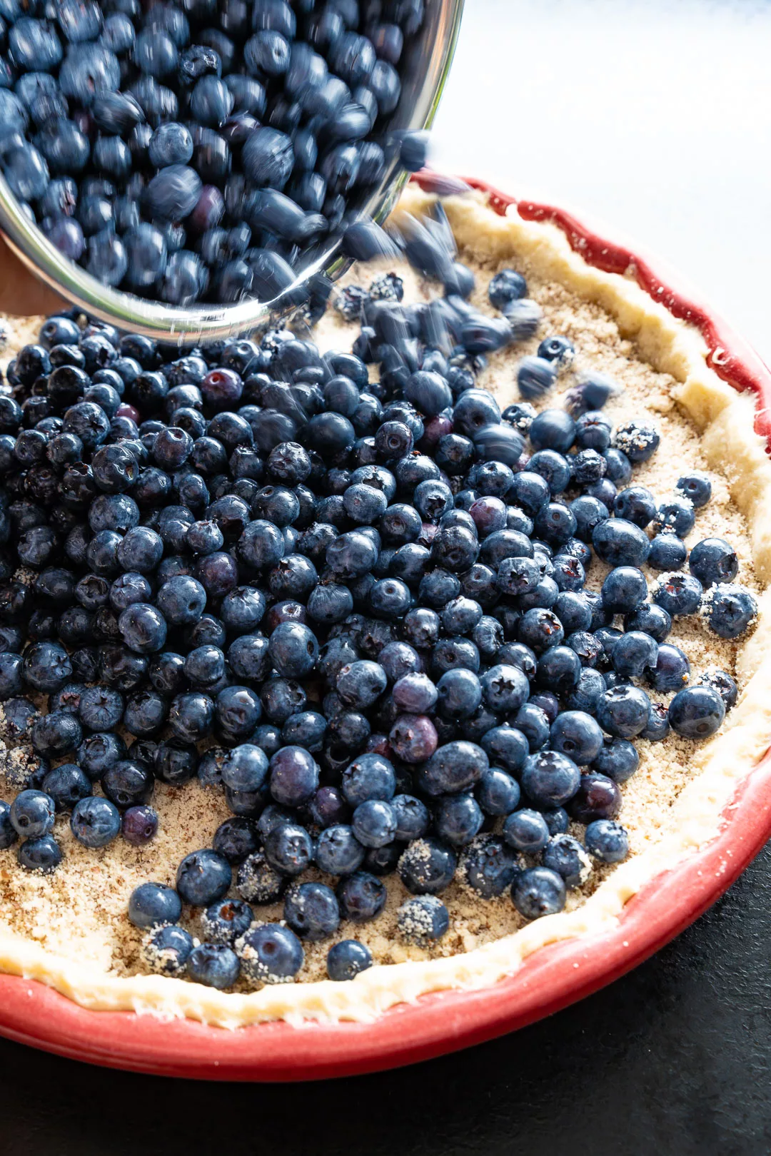 Frische Blaubeeren werden aus einer Schüssel auf einen vorbereiteten Tarteboden in einer roten Backform geschüttet.