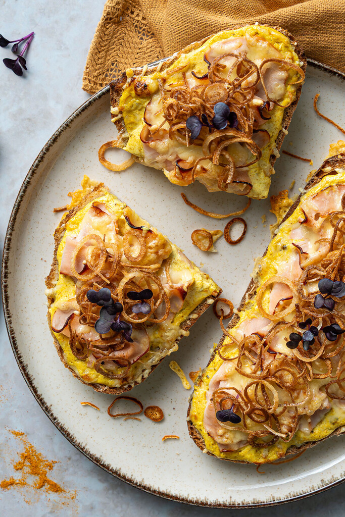 Herzhaft belegte goldene Stulle mit Hähnchen, Curry-Creme und knusprigen Röstzwiebeln auf einem rustikalen Teller, garniert mit frischen Microgreens.