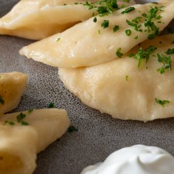Gekochte Wareniki mit Kartoffelfüllung auf einem Teller angerichtet, mit frischen Kräutern bestreut und serviert mit einem Klecks Sauerrahm