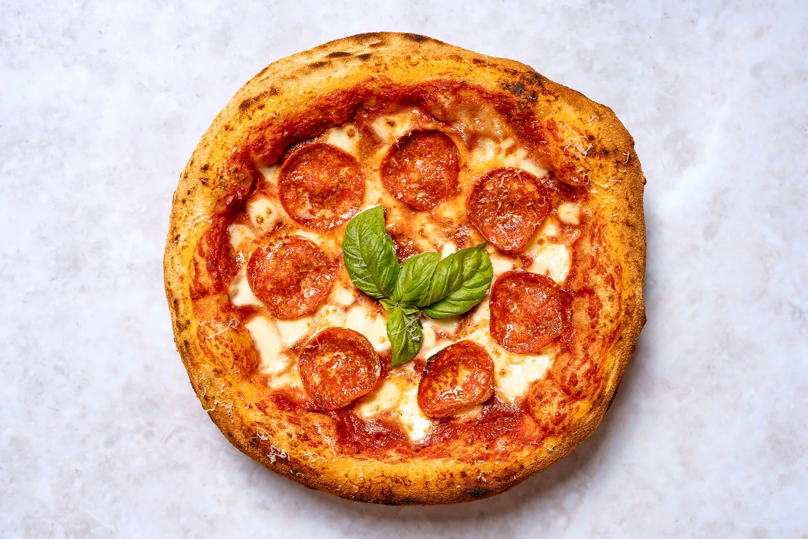 Draufsicht auf eine ganze, frisch gebackene Pizza Salami auf einem hellen Marmoruntergrund. Die Pizza hat einen hohen, luftig aufgegangenen und leicht leopardierten (punktuell dunkel gebräunten) Rand. Sie ist belegt mit roter Tomatensauce, geschmolzenem Mozzarella und mehreren Scheiben würziger Salami. In der Mitte der Pizza liegen als Garnitur frische, grüne Basilikumblätter.