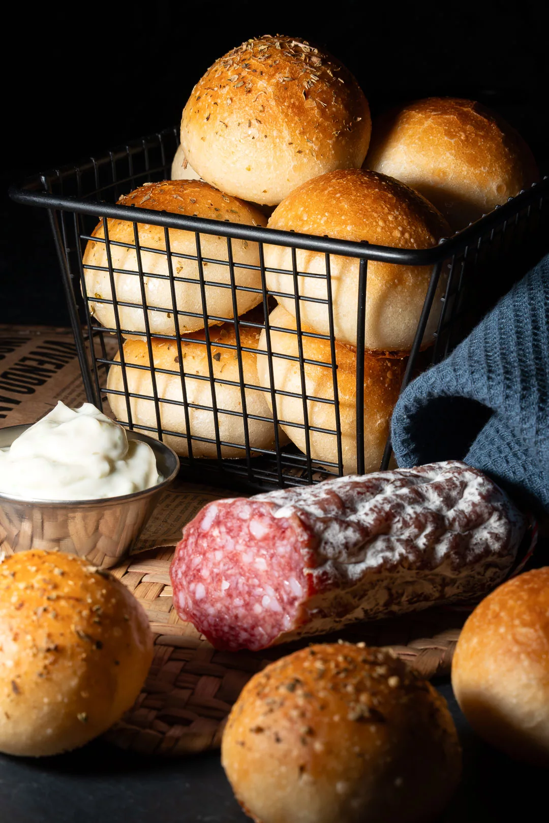 Rustikales Food-Foto von mehreren goldbraun gebackenen Pizzabrötchen, die teilweise in einem schwarzen Metalldrahtkorb und teilweise davor auf einem Flechtuntersatz liegen. Im Vordergrund liegt eine angeschnittene Salami mit Edelschimmelhülle und ein kleines Metallschälchen mit weißer Aioli. Die Szene ist in warmem, kontrastreichem Licht vor dunklem Hintergrund gesetzt, ergänzt durch ein blaues Küchentuch und Zeitungspapier im Vintage-Stil.