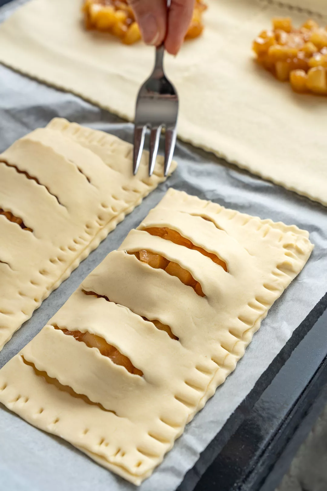 Vorbereitung von Apfeltaschen aus hellem Teig auf einem Backblech. Eine Person drückt den Rand einer Teigtasche mit einer Gabel fest. Die Taschen haben dekorative Schlitze, durch die die Apfelfüllung leicht zu sehen ist.