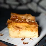 Ein Stück selbstgemachtes Galaktoboureko mit goldbraunem, knusprigem Filoteig und cremiger Grießfüllung, garniert mit Nüssen auf einem weißen Teller, umgeben von Zimtstangen und Kaffeebohnen.