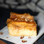 Ein Stück selbstgemachtes Galaktoboureko mit goldbraunem, knusprigem Filoteig und cremiger Grießfüllung, garniert mit Nüssen auf einem weißen Teller, umgeben von Zimtstangen und Kaffeebohnen.