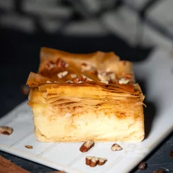 Galaktoboureko Rezept – griechisches Dessert mit Filoteig Ein Stück selbstgemachtes Galaktoboureko mit goldbraunem, knusprigem Filoteig und cremiger Grießfüllung, garniert mit Nüssen auf einem weißen Teller, umgeben von Zimtstangen und Kaffeebohnen.