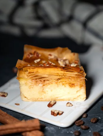 Ein Stück selbstgemachtes Galaktoboureko mit goldbraunem, knusprigem Filoteig und cremiger Grießfüllung, garniert mit Nüssen auf einem weißen Teller, umgeben von Zimtstangen und Kaffeebohnen.