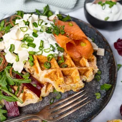 Herzhafte Kartoffelwaffeln mit Räucherlachs, Speck & Schnittlauchquark Knusprige Kartoffelwaffeln mit Speck, Räucherlachs, cremigem Schnittlauchquark und Frühlingszwiebeln auf einem Teller angerichtet, serviert mit frischem Salat und Dip.