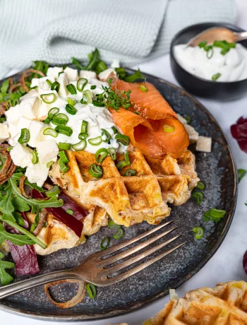 Knusprige Kartoffelwaffeln mit Speck, Räucherlachs, cremigem Schnittlauchquark und Frühlingszwiebeln auf einem Teller angerichtet, serviert mit frischem Salat und Dip.