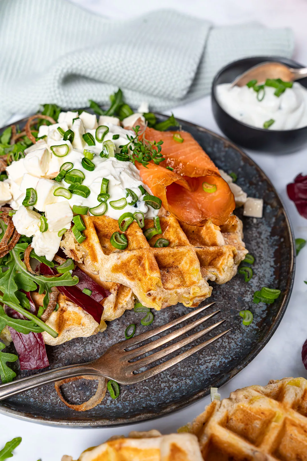 Knusprige Kartoffelwaffeln mit Speck, Räucherlachs, cremigem Schnittlauchquark und Frühlingszwiebeln auf einem Teller angerichtet, serviert mit frischem Salat und Dip.