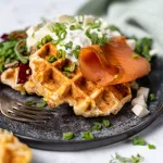 Knusprige Kartoffelwaffeln mit Räucherlachs, Speck, cremigem Schnittlauchquark und Frühlingszwiebeln auf einem dunklen Teller, garniert mit frischem Salat und serviert mit einer Gabel.