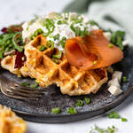 Knusprige Kartoffelwaffeln mit Räucherlachs, Speck, cremigem Schnittlauchquark und Frühlingszwiebeln auf einem dunklen Teller, garniert mit frischem Salat und serviert mit einer Gabel.