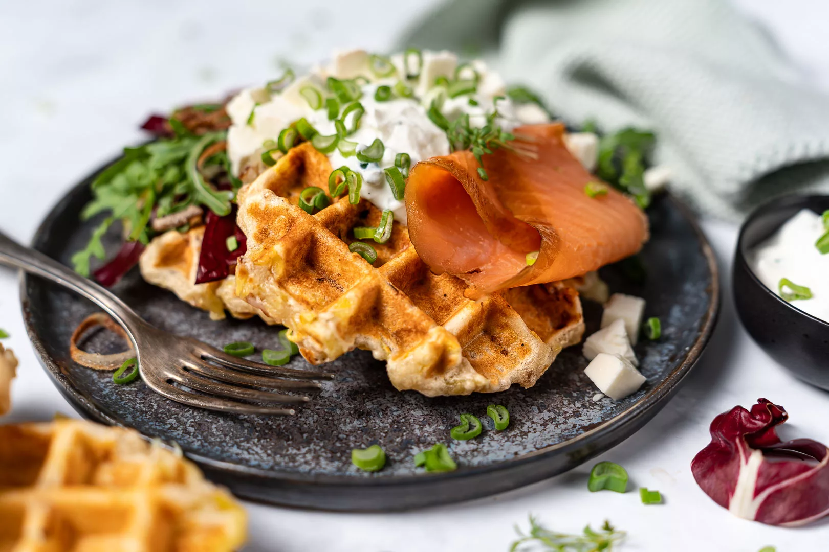 Knusprige Kartoffelwaffeln mit Räucherlachs, Speck, cremigem Schnittlauchquark und Frühlingszwiebeln auf einem dunklen Teller, garniert mit frischem Salat und serviert mit einer Gabel.