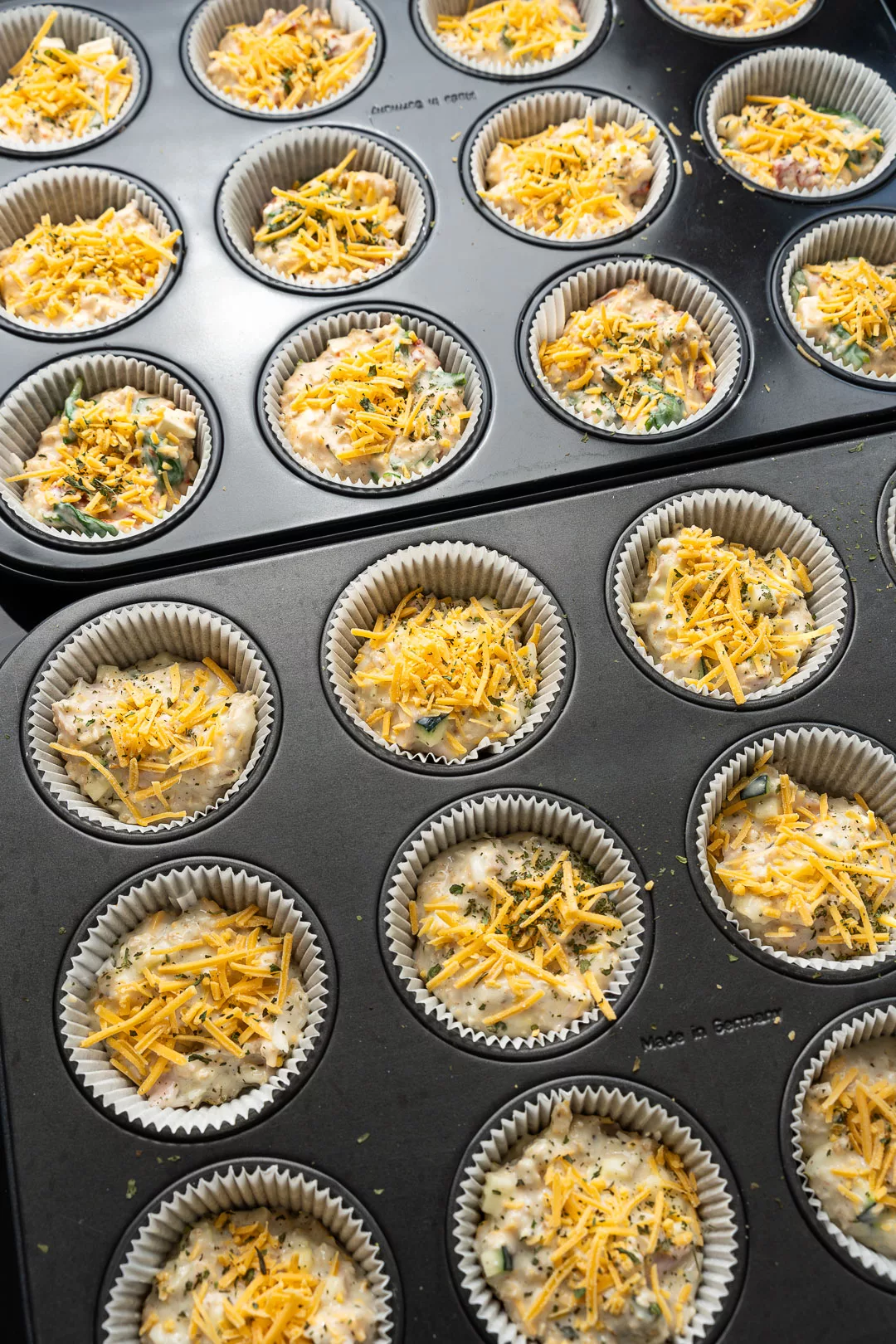 Muffinblech mit ungebackenen herzhaften Protein Muffins in Papierförmchen, gefüllt mit Teig, Gemüse und bestreut mit geriebenem Käse und Kräutern.