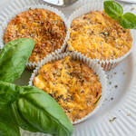 Drei gebackene herzhafte Protein Muffins mit Käse und Kräutern in Papierförmchen, serviert auf einem weißen Teller mit frischem Basilikum und einer Gabel im Hintergrund.