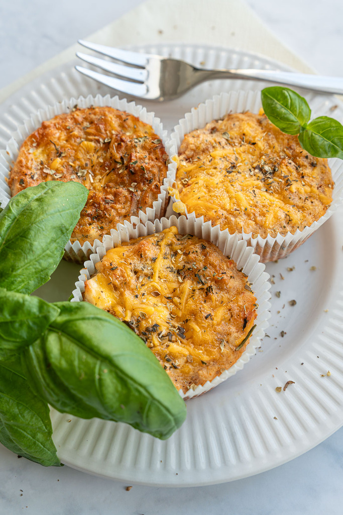 Drei gebackene herzhafte Protein Muffins mit Käse und Kräutern in Papierförmchen, serviert auf einem weißen Teller mit frischem Basilikum und einer Gabel im Hintergrund.