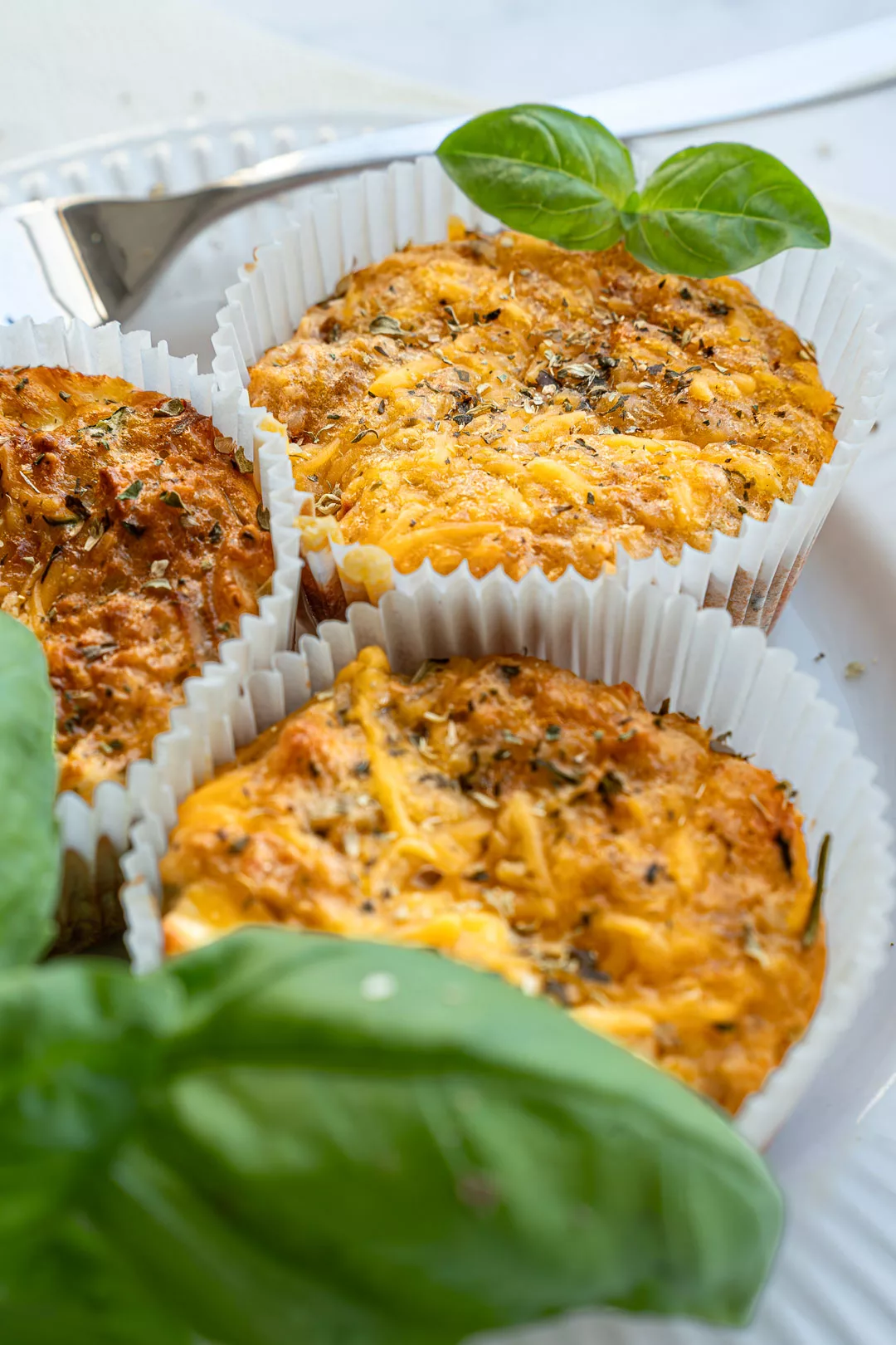 Nahaufnahme von drei goldbraun gebackenen herzhaften Protein Muffins mit Käse und Kräutern in Papierförmchen, garniert mit frischem Basilikum auf einem weißen Teller.