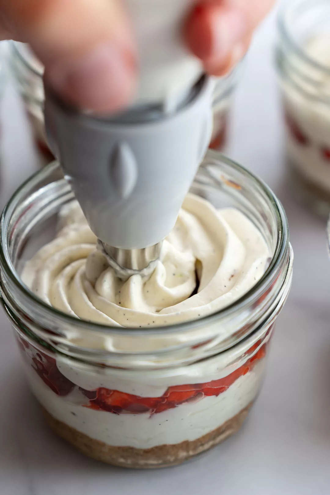 Mascarpone-Quark-Creme wird mit einem Spritzbeutel in ein Glas mit Keksboden und Erdbeerschicht für Erdbeer-Tiramisu gefüllt.