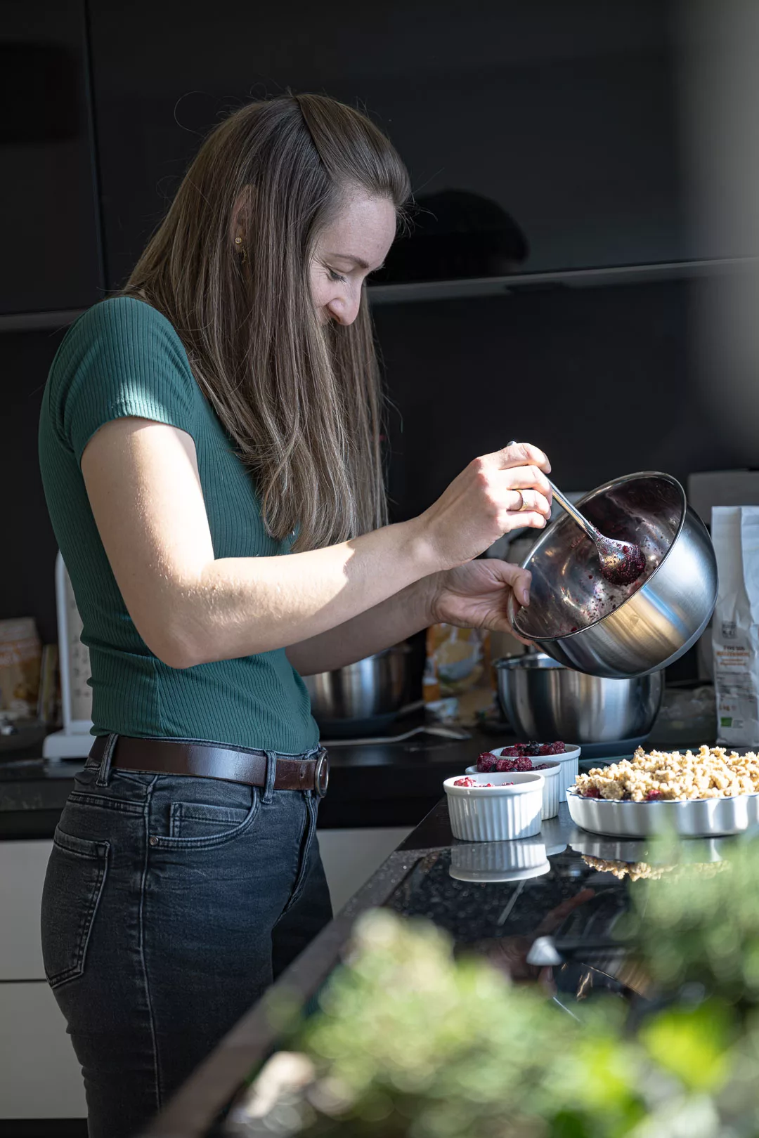 Eine Frau verteilt eine dunkle Beerenmischung aus einer Metallschüssel auf kleine weiße Förmchen; daneben steht eine große Form mit Beeren-Crumble und goldbraunen Streuseln.