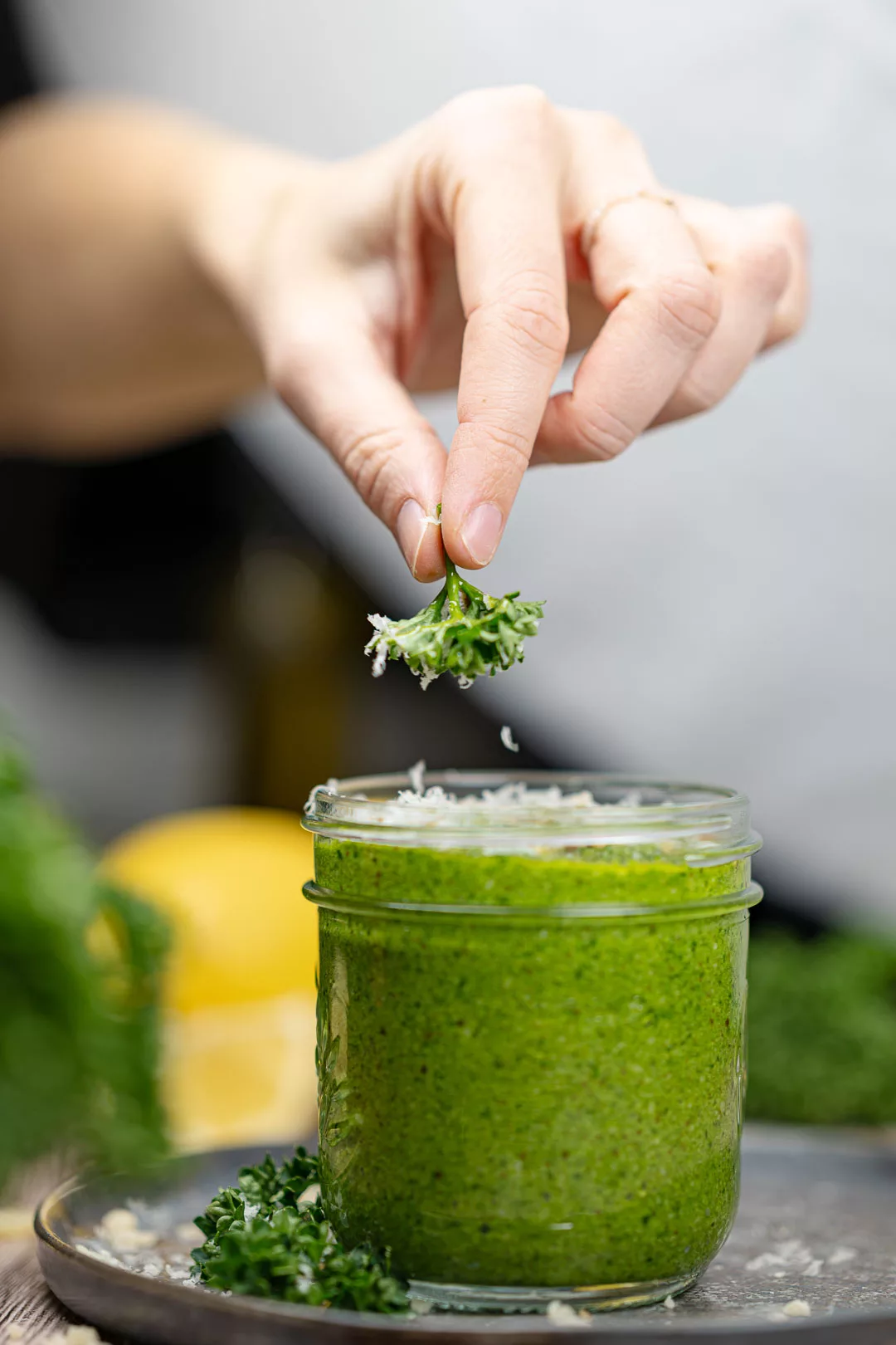 Hand garniert ein Glas mit cremigem grünem Kräuterpesto mit frischen Kräutern und geriebenem Käse; im Hintergrund liegt eine Zitrone.