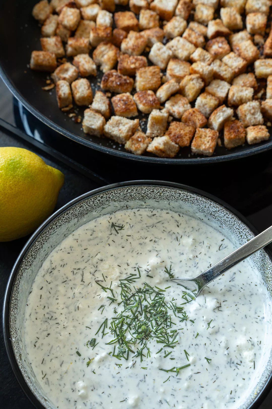 Schüssel mit cremigem Dill-Dressing und frischem Dill, daneben eine Zitrone und goldbraun geröstete Croutons in einer Pfanne.