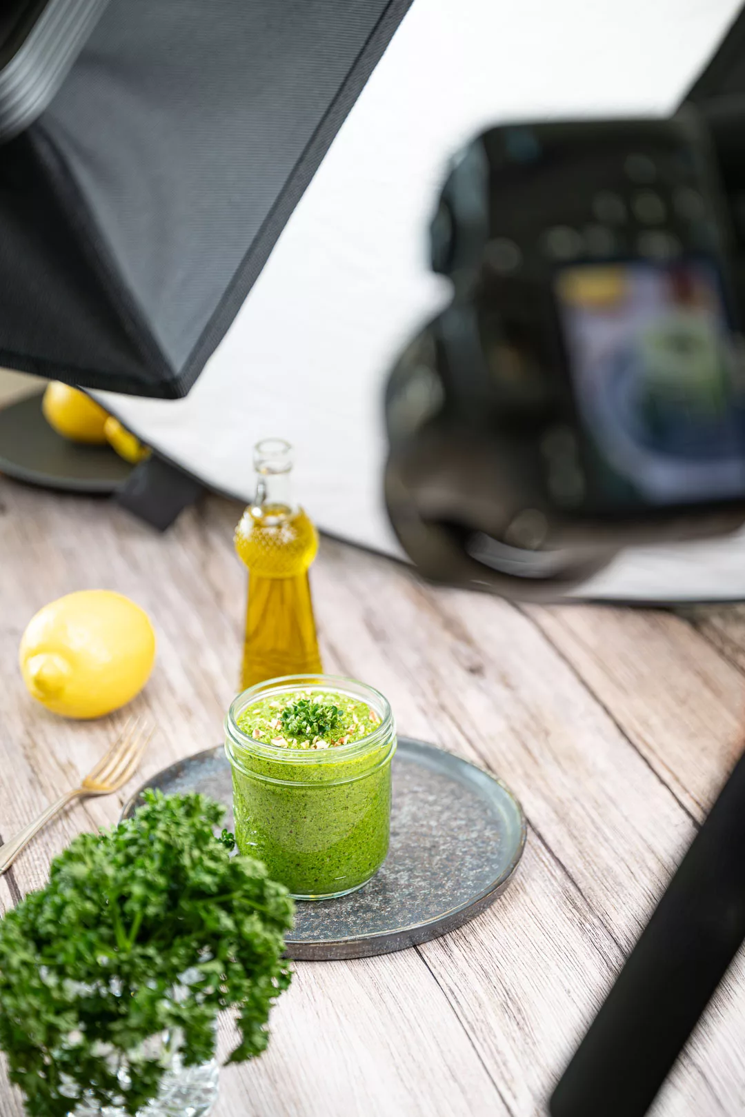 Foodfotografie-Setup mit Kamera, Softbox und Reflektor vor einem Glas grünem Pesto, umgeben von Petersilie, Zitrone und Olivenöl.