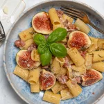 Teller mit Pasta, gebratenen Salsiccia-Stücken, frischen Feigenvierteln und Kräutern garniert.