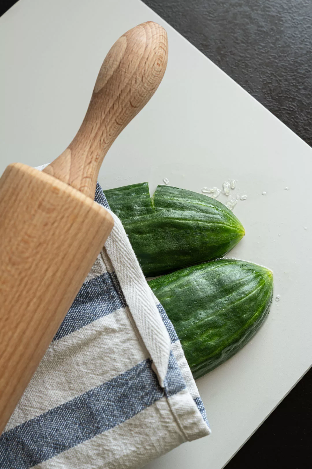 Zwei Stücke einer frischen Gurke auf einem weißen Schneidebrett neben einem Holzwerkzeug und einem blau-weiß gestreiften Küchentuch.