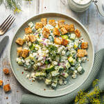 Smashed Gurkensalat mit Feta, roten Zwiebeln, frischem Dill und knusprigen Croutons auf einem grünen Teller, daneben ein Glas Kräuterdressing.