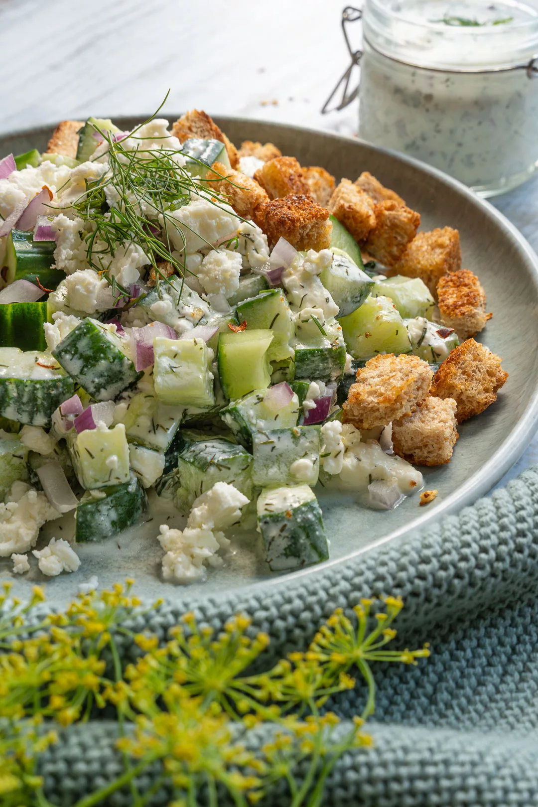 Nahaufnahme eines Smashed Gurkensalats mit Feta, roten Zwiebeln, frischem Dill und goldbraunen Croutons; im Hintergrund steht ein Glas Kräuterdressing.